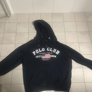 Svart hoodie från Polo Club - Säljer en svart hoodie från Polo Club med tryck på framsidan. Den har en klassisk känguruficka och justerbar huva med dragsko. Perfekt för en avslappnad stil.