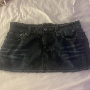 Mörkblå jeanskjol - Säljer en snygg mörkblå jeanskjol med klassisk femficksdesign och knappstängning fram. Perfekt för en avslappnad stil. Kjolen är kort och har en lätt slitning för en trendig look.