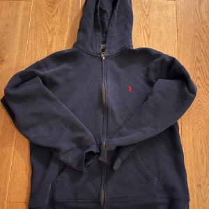 Mörkblå hoodie från Ralph Lauren - Säljer en snygg mörkblå hoodie från Ralph Lauren med dragkedja och det klassiska röda logotypbroderiet på bröstet. Perfekt för en avslappnad stil. Hoodien har långa ärmar och en praktisk ficka framtill.