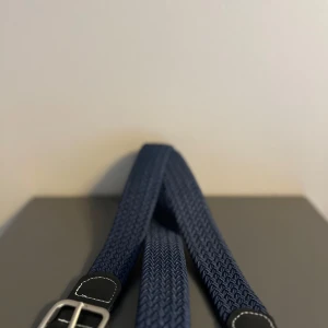 Blått flätat bälte - Snyggt blått flätat bälte med silverfärgat spänne. Perfekt för att ge en stilren touch till din outfit. Bältet har svarta läderdetaljer vid spännet som ger en extra dimension till designen.