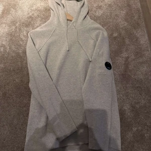 Grå hoodie från C.P. Company - Säljer en stilren grå stickad hoodie från C.P. Company. Den har en klassisk design med dragsko i huvan och en rund detalj på ärmen. Perfekt för en avslappnad look. 