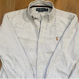 Randig skjorta från Ralph Lauren - Säljer en stilren vit och blå randig skjorta från Ralph Lauren. Skjortan har långa ärmar och en klassisk krage med knappar. Den är broderad med den ikoniska loggan på bröstet. Perfekt för en snygg och avslappnad look.