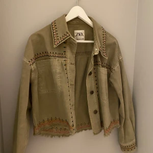 grön jacka från Zara - Säljer en olivgrön jacka från Zara med snygga broderade detaljer i flera färger. Jackan har en croppad passform och långa ärmar. Perfekt för att ge din outfit en unik touch!