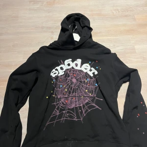 Svart hoodie Sp5der - Säljer en svart hoodie från Sp5der med en cool spindelnätsdesign och färgglada stjärnor på framsidan. Perfekt för en avslappnad stil med en unik touch. Hoodien har långa ärmar och en bekväm passform. Svart sp5der hoodie 