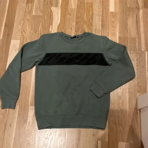 Grön tröja från Sail Racing - Säljer en stilren grön tröja från Sail Racing med en svart mesh-detalj över bröstet. Tröjan har långa ärmar och är rund halsad . Perfekt för en avslappnad stil.