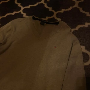 Grå tröja från GANT - Säljer en stilren grå tröja från GANT med v-ringning. Tröjan har en liten broderad logotyp på bröstet och är perfekt för en avslappnad look. Storlek XL men passar mer L och M