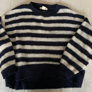 Randig stickad tröja - Mysig stickad tröja med breda marinblå och vita ränder. Tröjan har en avslappnad passform och långa ärmar. Den är gjord i ylle material som håller dig varm och bekväm.