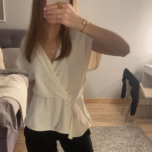 Vit omlottopp från Zara - Säljer en stilren vit omlottopp från Zara. Toppen har en elegant knytning i midjan och är ärmlös, vilket ger en luftig och modern look. Perfekt för att klä upp eller ner beroende på tillfälle.