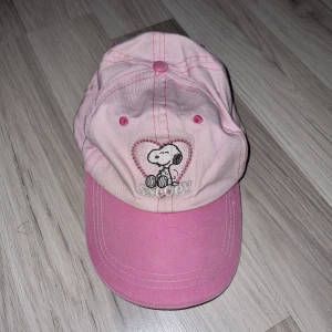 Rosa keps med Snoopy-motiv - Söt rosa keps med broderat Snoopy-motiv i ett hjärta på framsidan. Kepsen har justerbar rem baktill och är perfekt för en avslappnad stil. Tillverkad i bomull för en bekväm passform. Passar barn I ålder 5-8 år.