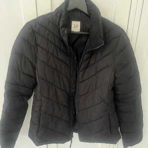 Svart quiltad jacka från GAP - Säljer en svart quiltad jacka från GAP. Jackan har en dragkedja framtill och en hög krage för extra värme. Perfekt för kyligare dagar och har en stilren design som passar till det mesta.