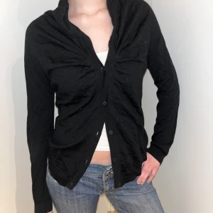 Svart kofta från COS - Säljer en stilren svart kofta från COS. Den har en klassisk design med knappar framtill och långa ärmar. Lite kortar på baksidan. Perfekt för lager-på-lager och passar till många olika outfits. Har dock ett hål vid armhålan men går enkelt att laga! 