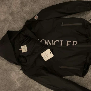 Svart vindjacka från Moncler - Snygg svart vindjacka från Moncler med huva och dragkedja. Jackan har Moncler-loggan på ärmen och ett stort tryck på framsidan. Perfekt för blåsiga dagar. Jackan är storlek M och har aldrig varit till användning. 
