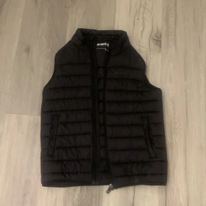 Svart dunväst från Everest - Säljer en svart dunväst från Everest. Västen har en dragkedja framtill och är perfekt för kyligare dagar. Den är stilren och lätt att matcha med olika outfits.