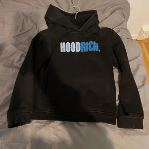 Svart hoodie från Hoodrich - Säljer en svart hoodie från Hoodrich med blå och vit text på bröstet. Den har en klassisk känguruficka och är tillverkad i en bomulls- och polyesterblandning. Perfekt för en avslappnad stil.
