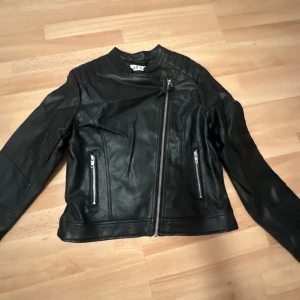 Svart skinnjacka från H&M - Snygg svart skinnjacka från H&M med asymmetrisk dragkedja och två framfickor med dragkedjor. Jackan har en stilren design med quiltade detaljer på axlarna och långa ärmar. Perfekt för en cool look!