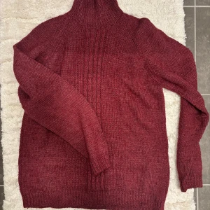 Vinröd stickad polotröja - Säljer en mysig vinröd stickad polotröja med långa ärmar. Perfekt för kyliga dagar med sin höga krage och ribbade detaljer. Passar utmärkt till både jeans och kjol. Hör av dig vid intresse!