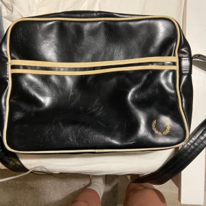 Svart axelväska från Fred Perry - Snygg svart axelväska från Fred Perry med beige detaljer och dragkedja. Väskan har en klassisk design med Fred Perry-loggan i hörnet. Perfekt för att bära dina dagliga nödvändigheter med stil.