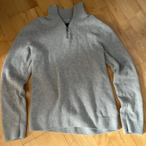 Grå tröja med dragkedja från Jack & Jones - Säljer en stilren grå tröja från Jack & Jones med dragkedja vid halsen. Perfekt för kyligare dagar med sin mjuka och bekväma passform. Tröjan har långa ärmar och en hög krage som ger extra värme.