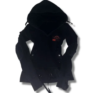 Svart hoodie med Volbeat-tryck - Snygg svart hoodie med Volbeat-tryck i rött. Den har hål i ärmarna där man kan placera tummarna (ska vara så). V-ring. Det finns även dekorativa knappar. Inga defekter förutom lite slitna vid ärmarnas slut. Skriv om du har några funderingar!