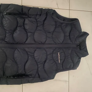 Mörkblå dunväst från Peak Performance - Snygg mörkblå dunväst från Peak Performance med dragkedja och diskret logga på bröstet. Perfekt för kyliga dagar med sin quiltade design som håller värmen. Västen har en stilren look och passar bra till olika outfits.