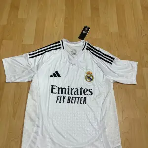 Säljer en vit Real Madrid fotbollströja från Adidas med korta ärmar och svarta detaljer. Tröjan har klubbens emblem på bröstet och texten 'Emirates Fly Better' på framsidan. Perfekt för alla fotbollsfans!