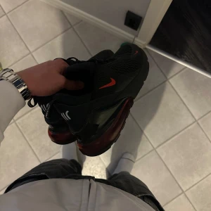 Svarta Nike Air Max 270 - Snygga svarta Nike Air Max 270 med röda detaljer. Skorna har en stor synlig Air-enhet i hälen för extra komfort och stil. Perfekta för både träning och vardag. De har en modern design med snörning och Nike-logga på sidan. INTE ANVÄNDA, passar 43,44