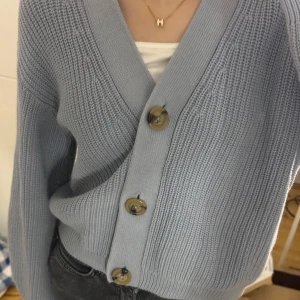 Ljusblå stickad kofta från H&M - Säljer en ljusblå stickad kofta från H&M med stora knappar framtill. Koftan har en v-ringad design och långa ärmar, perfekt för kyligare dagar. Enkel och stilren, passar till många olika outfits.