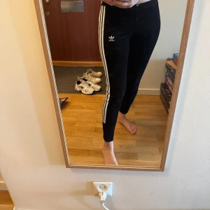 Svarta Adidas leggings med vita ränder i mocka  - Säljer ett par svarta Adidas leggings med klassiska vita ränder längs sidorna och Adidas-logga på låret. Tight passform och elastiskt material som sitter skönt. Perfekta för träning eller en sportig vardagslook.