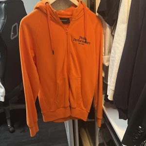 Orange hoodie från Peak Performance - Säljer en orange hoodie från Peak Performance med dragkedja och huva. Tröjan har fickor framtill och svart broderad logga på bröstet. Perfekt för dig som gillar färg och vill ha något bekvämt och snyggt.
