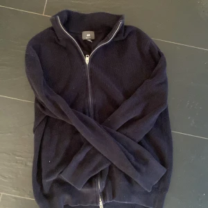 Mörkblå stickad kofta från H&M - Säljer en mörkblå stickad kofta från H&M med dragkedja framtill. Tröjan har lång ärm och hög krage, perfekt för kyliga dagar. Enkel och stilren design som passar till mycket.