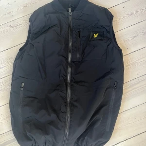 Svart väst från Lyle & Scott Junior - Säljer en svart väst från Lyle & Scott Junior med dragkedja framtill och två fickor med dragkedja. Västen har ett gult logomärke på bröstet och är perfekt för lager-på-lager-stil.