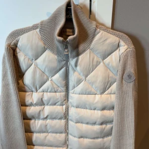 Moncler cardigan - Säljer en snygg beige quiltad jacka från Moncler med stickade ärmar och hög krage. Jackan har dragkedja framtill och Moncler-logga på ärmen. Perfekt för dig som vill ha en stilren och exklusiv look.‼️ OBS( storlek L passar S)