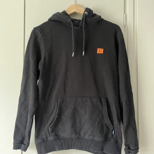 Svart hoodie med orange detalj - Svart hoodie från Les Deux med orange logga på bröstet. Tröjan har huva med dragsko och en stor ficka framtill. 