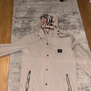 Beige Burberry jacka  - Säljer en beige jacka från Burberry med klassiskt rutigt foder i huvan. Jackan har dragkedja och knappar framtill samt två fickor med dragkedja. Helt ny