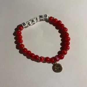 Rött pärlarmband med texten BE KIND - Armband med röda pärlor och vita bokstavspärlor som bildar texten 'BE KIND'. Liten rund berlock i guld hänger vid sidan. Perfekt accessoar för att sprida positivitet. Inga defekter. Köpte från indiska 🤍