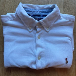 Vit kortärmad skjorta från Polo Ralph Lauren - Klassisk vit kortärmad skjorta från Polo Ralph Lauren med button-down krage och broderad logga på bröstet. Skjortan har knappar framtill och är tillverkad i ett mjukt bomullsmaterial. Perfekt för en stilren och avslappnad look.