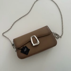 Beige axelväska från Rizzo med silverkedja - Säljer en stilren beige axelväska från Rizzo med silverfärgad kedja och spänne. Väskan har ett elegant och minimalistiskt utseende, med flera fack inuti och dragkedja för extra förvaring. Väskan är väldigt rymlig och perfekt för dig som gillar klassisk design. Den är helt ny och oanvänd med prislapp på.