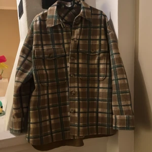 Rutig overshirt från Oysho - Säljer en rutig overshirt från Oysho i bruna, gröna och beige toner. Skjortan har två bröstfickor med lock, knäppning framtill och klassisk krage. Perfekt att slänga över en t-shirt för en avslappnad look.