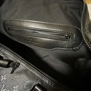 Svart weekendbag från Louis Vuitton - Säljer en lyxig svart weekendbag från Louis Vuitton med klassiskt monogrammönster i präglat skinn. Väskan har dubbla handtag, dragkedja och detaljer i svart läder. Perfekt för resor och har en rymlig insida med innerficka med dragkedja.