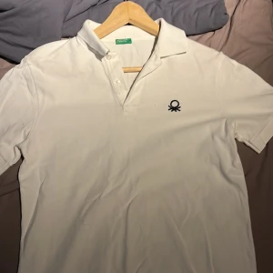 Vit pikétröja från United Colors of Benetton str 170 cm - Klassisk vit pikétröja från United Colors of Benetton med svart broderad logga på bröstet. Tröjan har krage och knappar framtill samt korta ärmar. Perfekt för en stilren och enkel look.