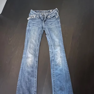 Blå broderade jeansbyxor från Miss Me - Säljer ett par blå jeansbyxor från Miss Me med snygga broderade detaljer och dekorativa knappar på bakfickorna. Byxorna har bootcut-modell och klassisk femficksdesign. Perfekta för dig som vill sticka ut med coola detaljer.