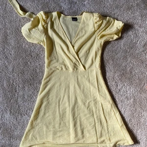 Ljusgul omlottklänning från Gina Tricot - Supersöt ljusgul omlottklänning från Gina Tricot med korta puffärmar och knyt i midjan. Klänningen har v-ringning och är tillverkad i ett mjukt bomullsmaterial. Perfekt för vår och sommar!🌼🌻