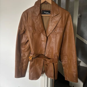 Brun skinnjacka med bälte - Säljer en brun skinnjacka från Genuine Leather med bälte i midjan och stora slag. Jackan har långa ärmar, knappar framtill och två bröstfickor. Perfekt för dig som vill ha en klassisk och stilren look.