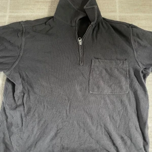 Halfzip från Arket - Mörkgrå tröja med halv dragkedja och hög krage. Tröjan har en bröstficka på vänster sida och är tillverkad i ett mjukt, stickat material. Perfekt för lager-på-lager och enkel att matcha med andra plagg.