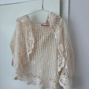 Beige virkad kofta med volangkant - Superfin beige virkad kofta med volangkant och blommiga detaljer. Koftan är luftig och har korta ärmar, luva och knäppning fram. Sitter bra på mig som brukar bära S/M. Tveka inte på att höra av dig vid frågor eller prisförslag!