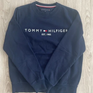 Mörkblå sweatshirt från Tommy Hilfiger - Säljer en mörkblå sweatshirt från Tommy Hilfiger med klassisk logga och texten 'EST. 1985' broderad på bröstet. Tröjan har rund halsringning och långa ärmar. Perfekt för en stilren och avslappnad look.