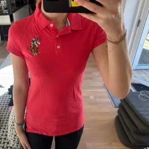 Röd pikétröja från US polo  - Säljer en klassisk röd pikétröja från us polo med broderad logga och polospelare på bröstet. Tröjan har korta ärmar, krage och knäppning framtill. 