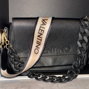 Svart crossbody väska från Valentino - Snygg svart axelväska från Valentino med bred justerbar axelrem i beige och svart med logga. Väskan har en stor kedjedetalj och guldiga metalldetaljer. Tillverkad i skinnliknande material och har dragkedja på insidan. Notera att det är Mario Valentino! Kom gärna med bud, kan sänka priset!