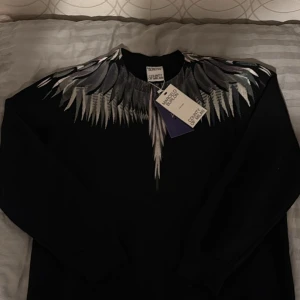  Marcelo Burlon - Säljer en svart sweatshirt från Marcelo Burlon med ikoniskt vingmönster i grått och vitt över axlar och bröst. Tröjan har rund hals och långa ärmar. Perfekt statement-plagg för dig som vill sticka ut. Helt ny 