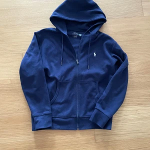 Zip hoodie från Ralph Lauren - Säljer en mörkblå hoodie från Polo Ralph Lauren med dragkedja och klassisk logga broderad på bröstet. Tröjan har huva, långa ärmar och två fickor framtill. Perfekt för en avslappnad stil.